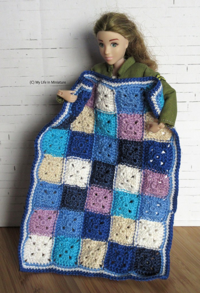 Sarah stands against a white brick background, holding a crochet blanket up in front of her. She wears her khaki jacket, and is looking down at the blanket. The blanket is in the same shades of blue, purple, cream, and white as the first one, but this one is made of single-coloured squares in a 7x5 rectangle. 