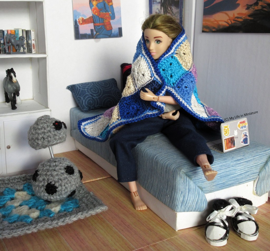 Sarah sits on her bed, one leg tucked underneath herself, with the blue blanket over her head and shoulders as a cape. She wears navy pants and a grey shirt, and her shoes are at the end of the bed. She smiles at Chip, who is on the floor beside the bed. Chip has their head raised in curiosity. 