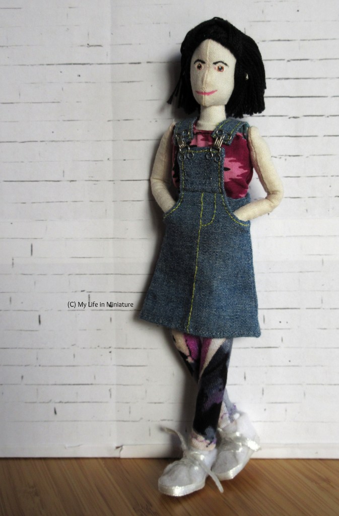 Tiffany wears the leggings with the same colourful shirt as earlier, this time with a denim pinafore over the top. She has her hands tucked into the pinafore's pockets and one foot tucked behind the other ankle.