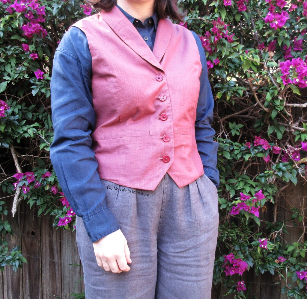 The author stands in front of a purple bougainvillea bush, photographed from neck to thighs. She wears a dusky pink collared waistcoat with a navy shirt and dark grey pants. She has one hand in her pant pocket, and the other hanging beside her. 