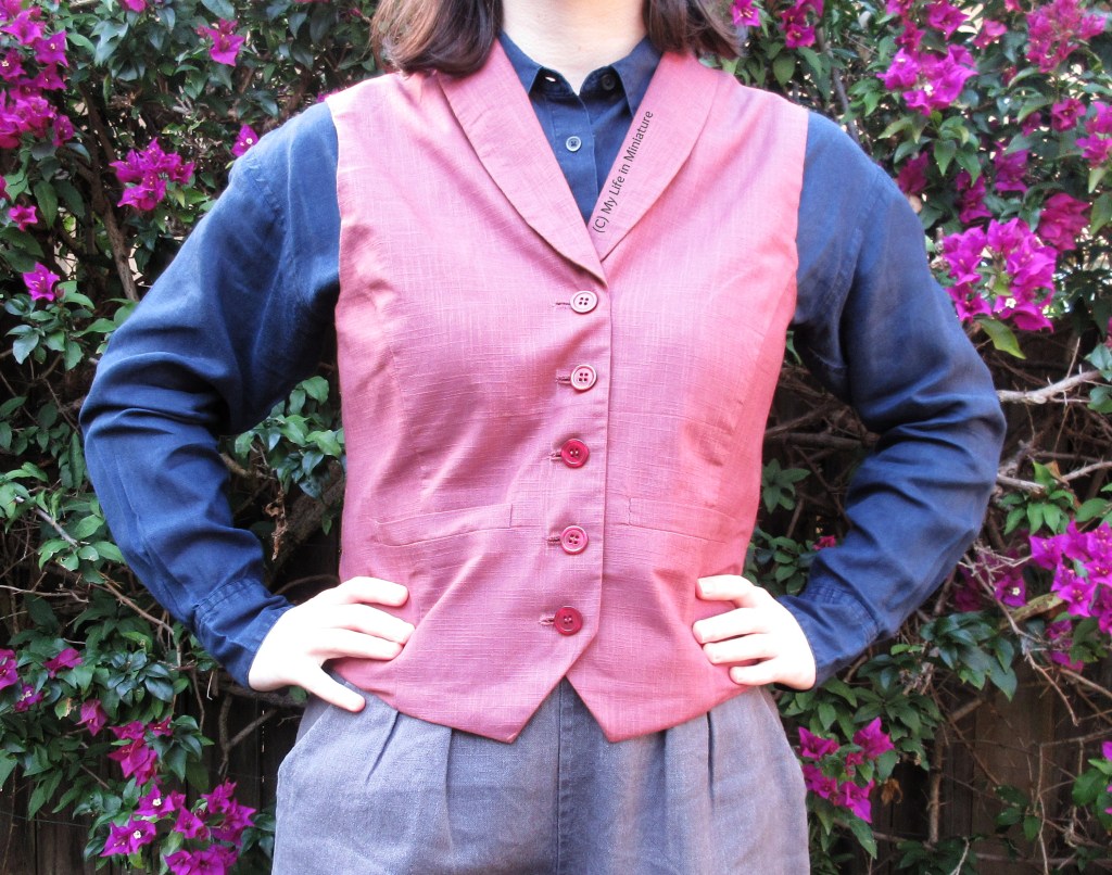 The author stands in front of a bougainvillea bush, wearing the pink waistcoat/navy shirt combo. She fully faces the camera, and has her hands on her hips. She is photographed from neck to thighs. 