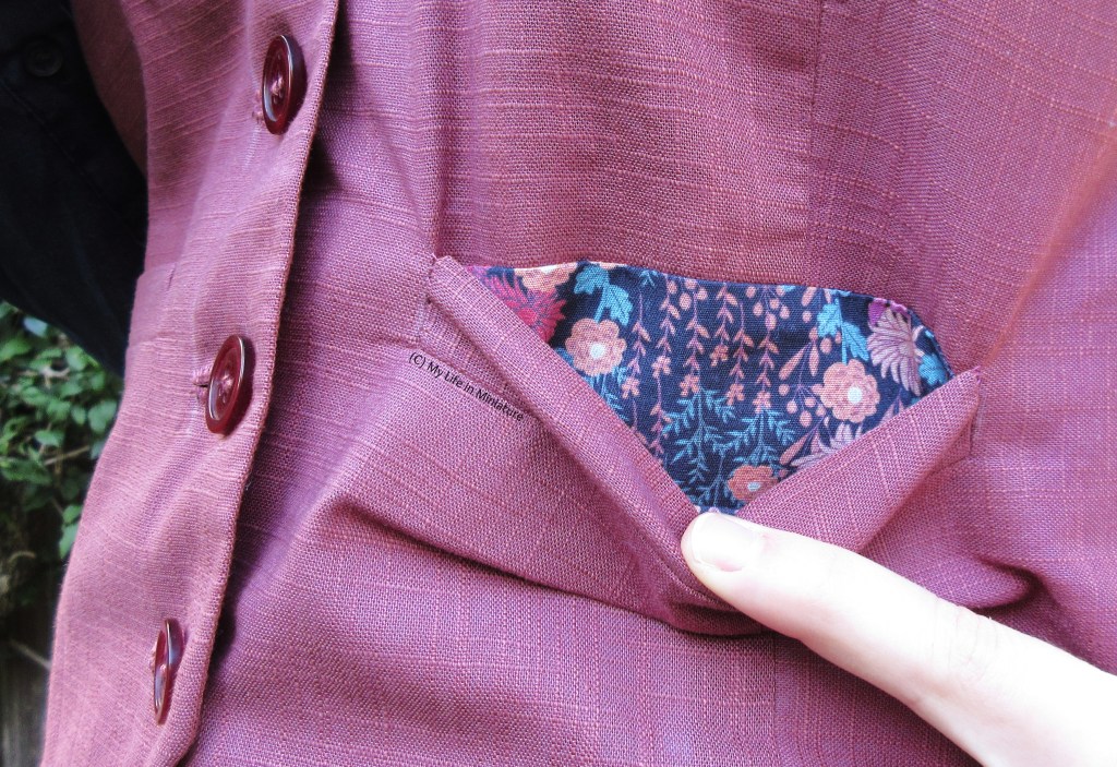 Close-up of one of the welt pockets. The author's hand is pulling the pocket open gently, exposing the lining. The lining is a pink floral fabric with pink and turquoise leaves on a navy background. 