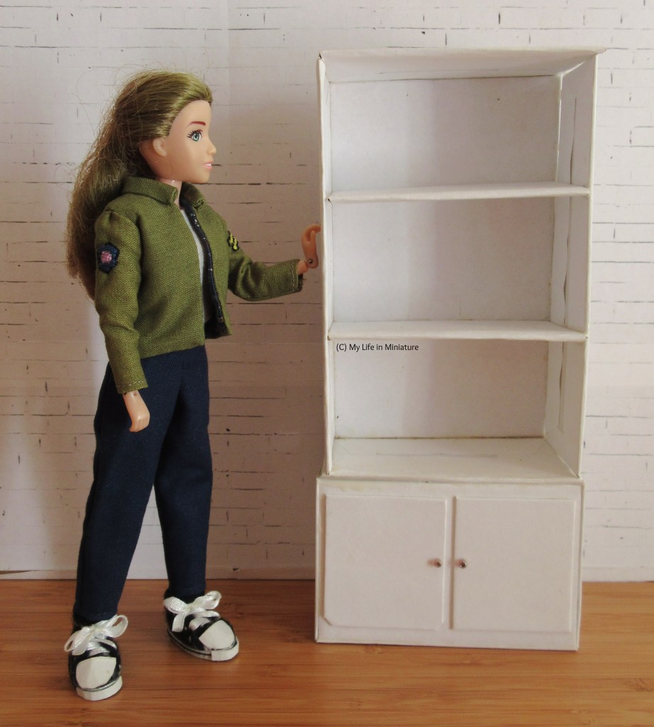 Sarah stands next to a white shelving unit. The shelving unit has a cupboard at the base with two doors. Above it are three shelves, including the top of the cupboard. Sarah wears a khaki jacket with navy pants and black shoes, and is facing the shelves. She has one hand resting flat on the side of the shelves. 