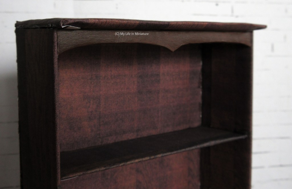 Close-up of the top shelf and the top of the bookcase. Underneath the top is an extra beam, shaped to be pointy in the middle and ends with soft curves between the points. It's been painted dark brown, a very close colour match to the wood. 