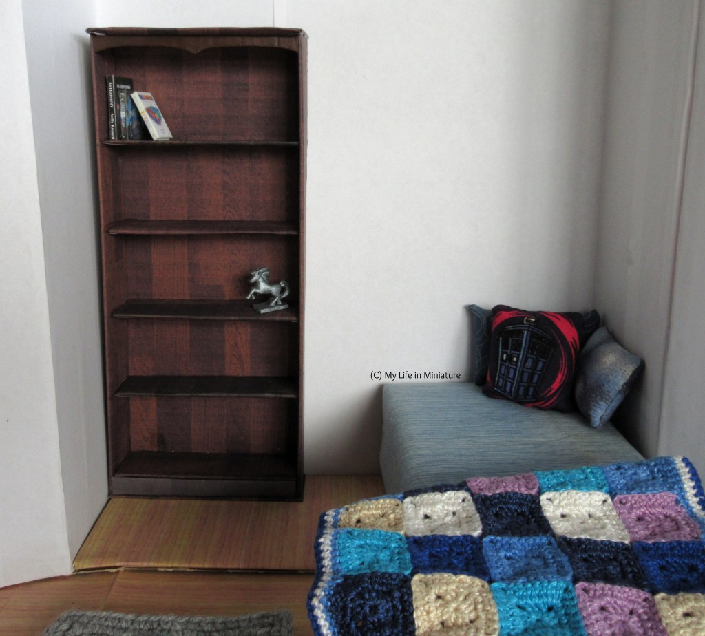 The bookshelf stands in the corner of Sarah's bedroom. It's in the same recess as Sarah's bed, in the opposite corner. On the shelves are a few books and a small silver horse statue. The bed has a few cushions on it, and a crocheted blanket at the foot. The walls of the room are white. 