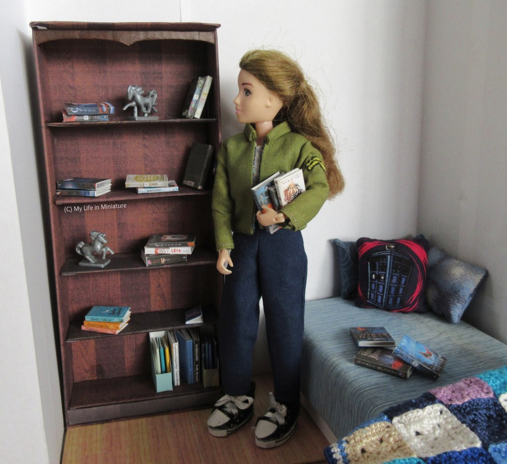 Sarah stands next to the bookshelf, with several books in her arms. She looks at the shelves, which are covered in stacks of her novels, a few textbooks and notebooks, and some silver horse statues. There are also a few books on the bed. 