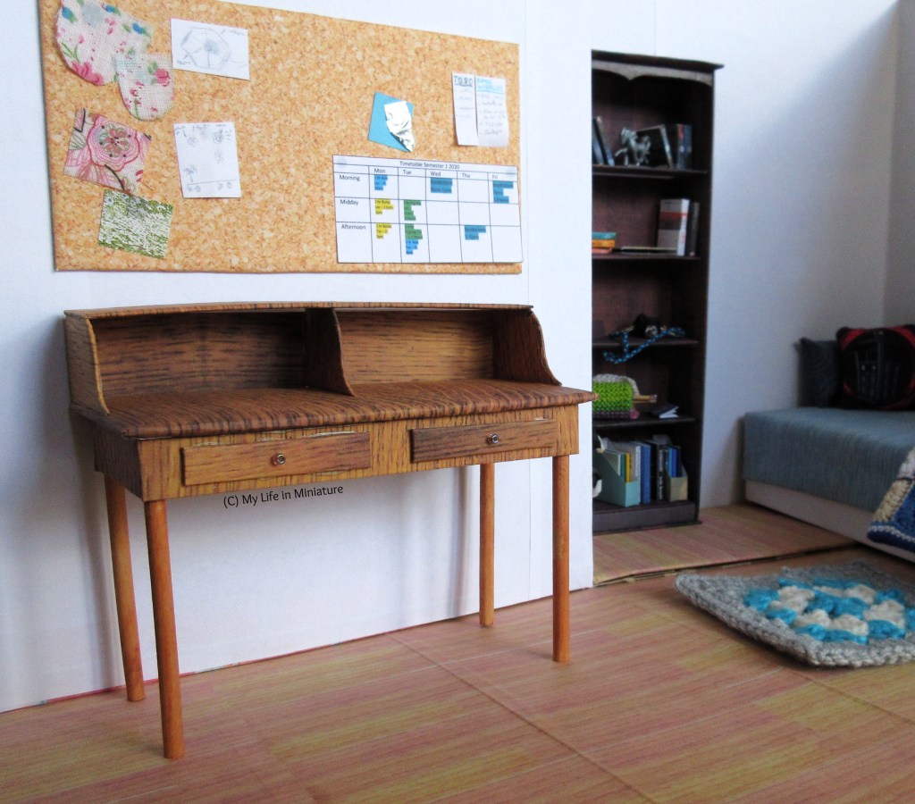 A wooden desk stands in Sarah's bedroom, underneath the corkboard. It is a lighter wood than the bookcase, which is visible in the background alongside the bed. The desk has an inbuilt shelf, resting on top of curved risers, and the space underneath the shelf is open for storage. The desk also has two slim drawers underneath the front of the work surface. 