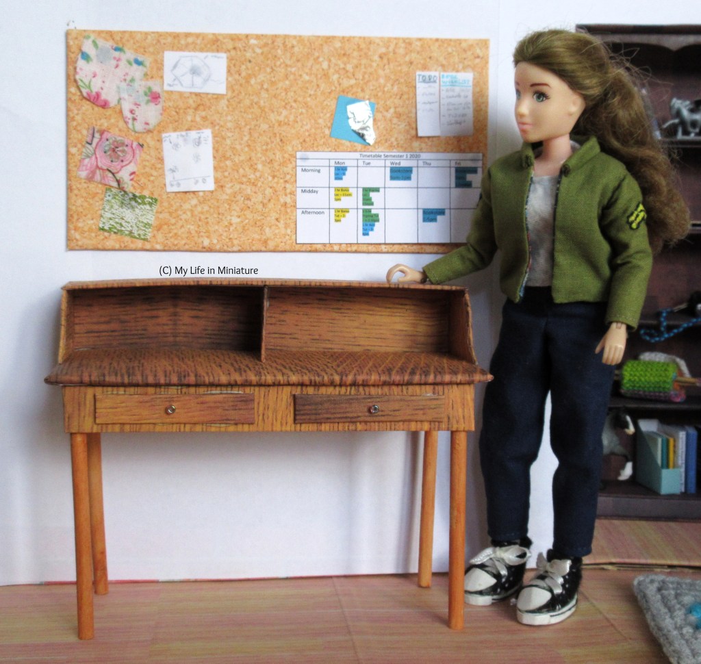 Sarah stands beside the new desk in her bedroom, one hand resting on the inbuilt shelf. She wears her khaki jacket with navy pants, a grey shirt, and black shoes. She is looking at the desk, thinking. 