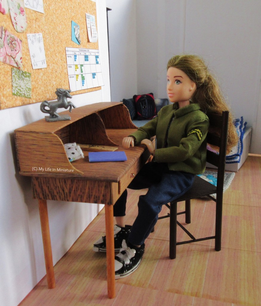 Sarah sits at the desk, angled slightly towards the camera. She has one elbow resting on the desk, and is looking at the silver horse statue. The notebook, ruler, and pencilcase are just in front of her on the desk's surface. Behind Sarah is her bed and the bedroom door. 
