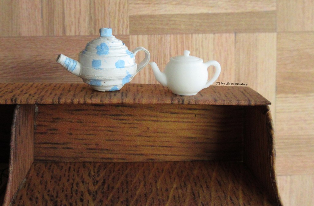 Two miniature teapots sit on the shelf of Sarah's new desk. The one on the left is white with pale blue spots painted on. It is clearly made of rolled paper, in that it is not perfectly spherical. The one on the right is smaller, also white, and looks a lot cleaner.