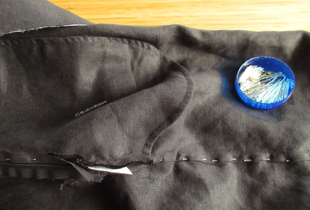 A closer-up photo of the pinned-down raw edges of the black sewing project. The end of the zip is visible, and between that and the pinned-down edges are a few edges still raw. In the top right is a blue pin tin, resting on the black fabric.