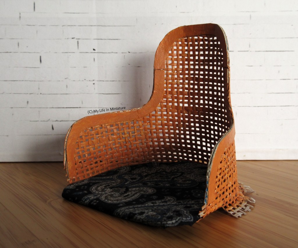 The seat of a chair is photographed against a white brick background. The chair back is red-brown lattice, with solid trim around the top edge. The seat is covered with blue fabric. The bottom lattice edge is in the process of being glued underneath the seat, so there are tabs splayed out around the edge of the seat.