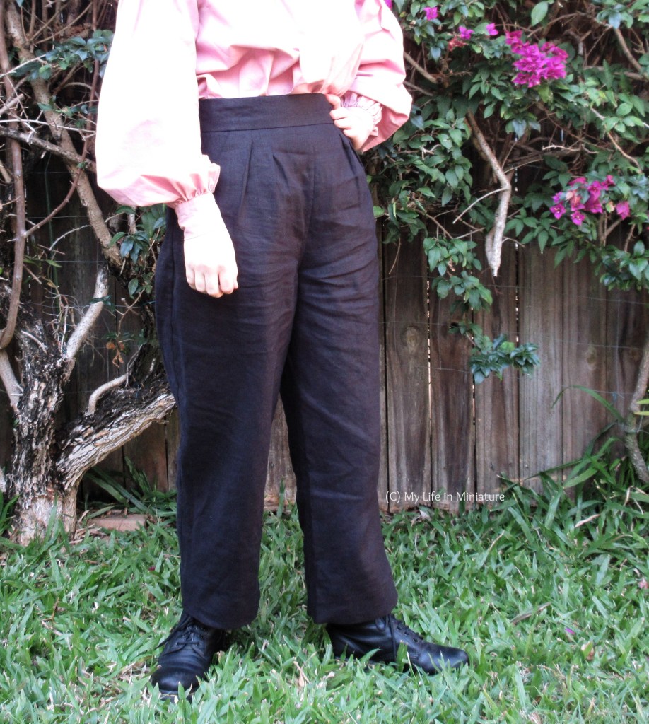 The author poses against a hedge, standing on grass. She wears black linen pants, a pink pirate shirt, and black boots. Her body is angled away from the camera, with her far hand on her hip and her near hand hanging beside her. 