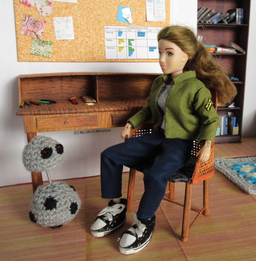 Sarah is mostly seated on a brown woven latticed chair, hands on the armrests as she lowers herself in. She's smiling as Chip the android watches on. Also visible in the scene is the desk, the corkboard above, and the bookshelf.