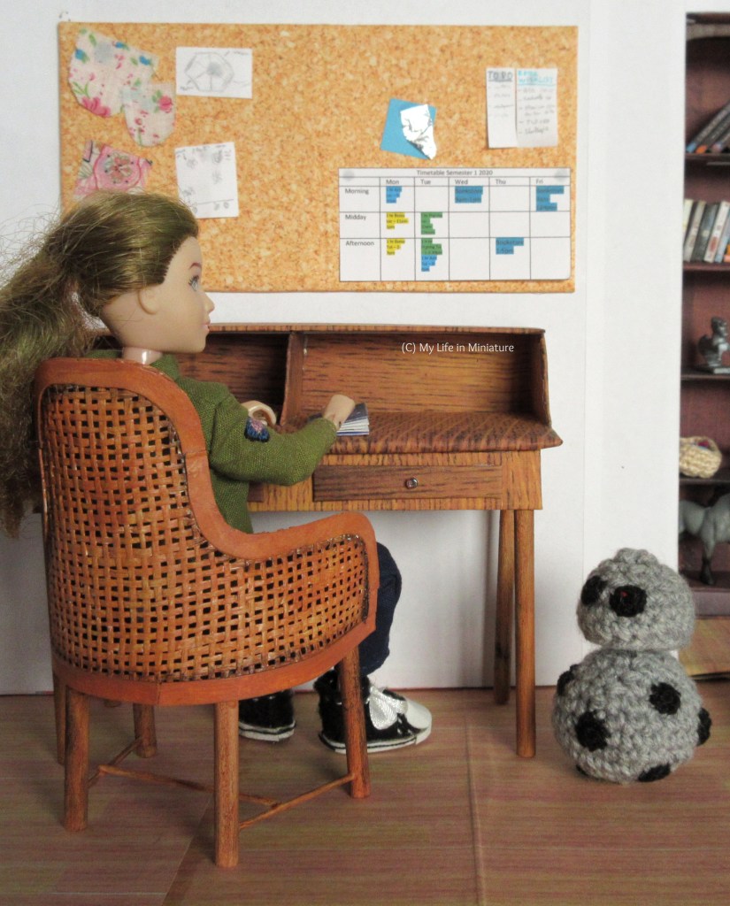 Sarah sits in the chair, back to the camera. She has a hand on a notebook on the desk, and is looking at Chip, who sits beside the desk. The back of the chair is more visible; one can see the lattice has grown darker around the edges where it meets the solid border. 