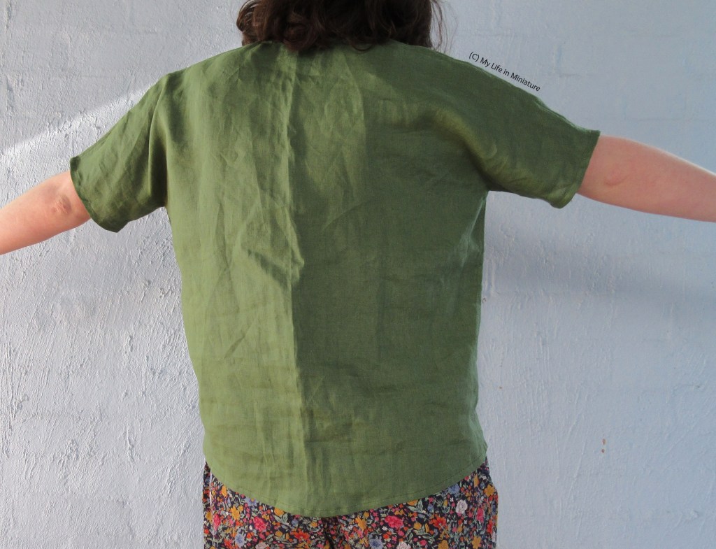 The author stands in front of a pale blue wall, photographed from shoulders to thighs. She faces away from the camera with arms outstretched. She wears the green linen shirt untucked from her pants, and the camera can see the plain back of the shirt.  