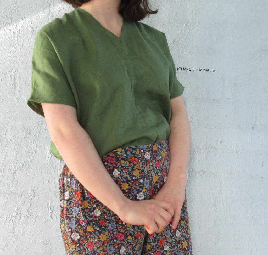 The author stands against a pale blue wall, photographed form shoulders to thighs. She wears a loose short-sleeved green linen shirt with floral cotton pants. She has her hands clasped in front of her, and her body is angled away from the camera slightly. 