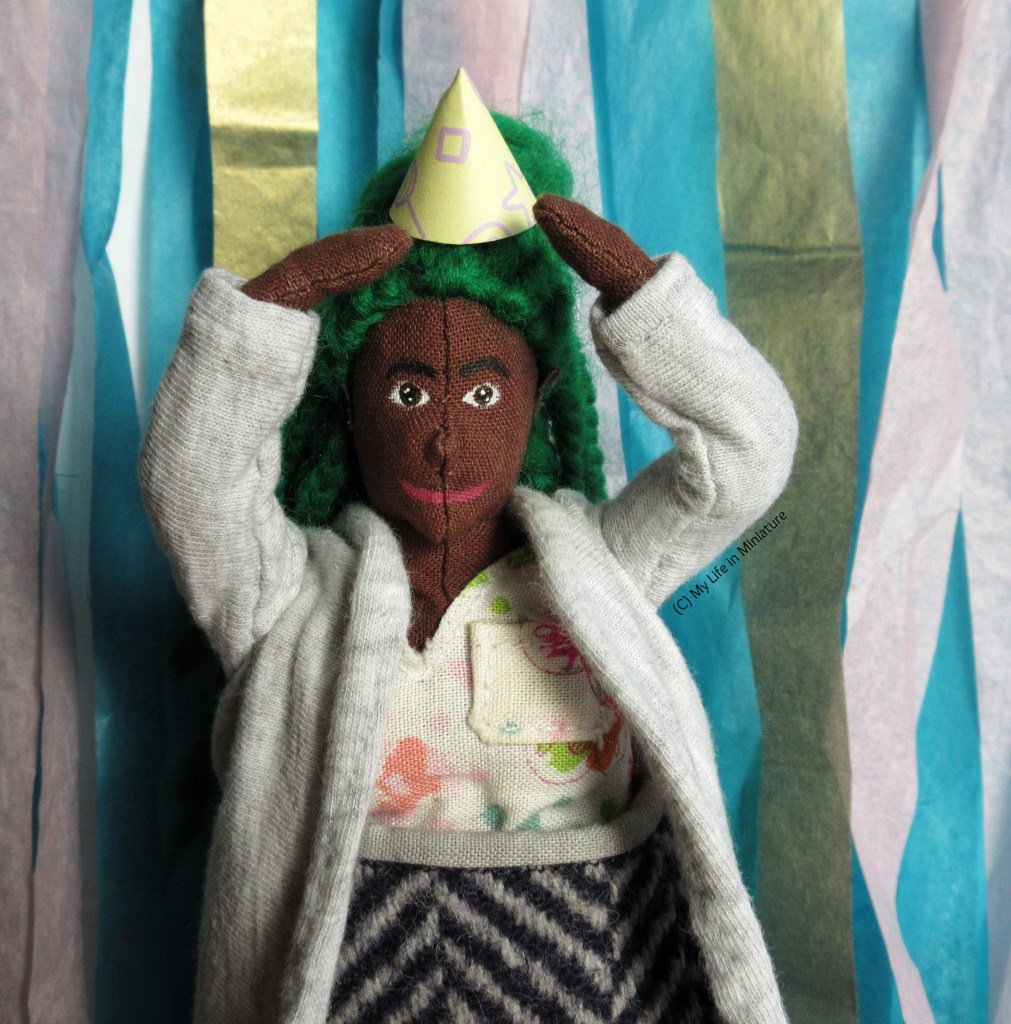 Hazel stands in front of a wall of streamers, holding a paper party hat on her head, just in front of her bun. She is smiling at the camera, and wears a grey cardigan, pink floral shirt, and a wool skirt. 