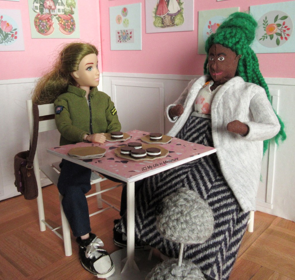 Sarah and Hazel sit at one of the tables in Fierro's, with a pastry each in front of them. Hazel is gesticulating as she explains something to Sarah, who is listening intently. Sarah wears her khaki jacket with navy pants and black shoes, and has her brown satchel hanging over the back of the chair. There are two extra small plates stacked next to Sarah on the table, beside the rest of the pastries. 