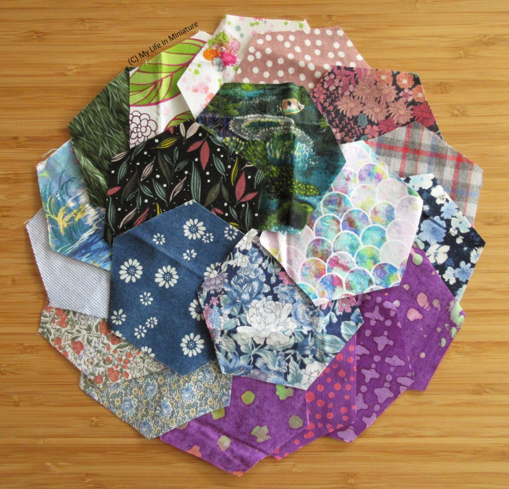 20 fabric hexagons are arranged on a wood surface in a circular fashion. Some of the patterns are more visible, in shades of pink, green, blue, and purple. There are stripes, florals, nautical prints, spots, and batik-dyed fabrics.