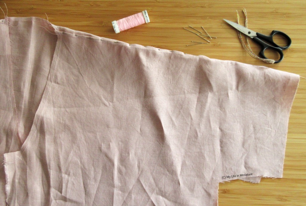 The neckline and sleeve of an unfinished linen shirt is laid flat on a wood surface. The shoulder seam has pins in it, and just above the seam is a thread spool, scissors, and pins. 
