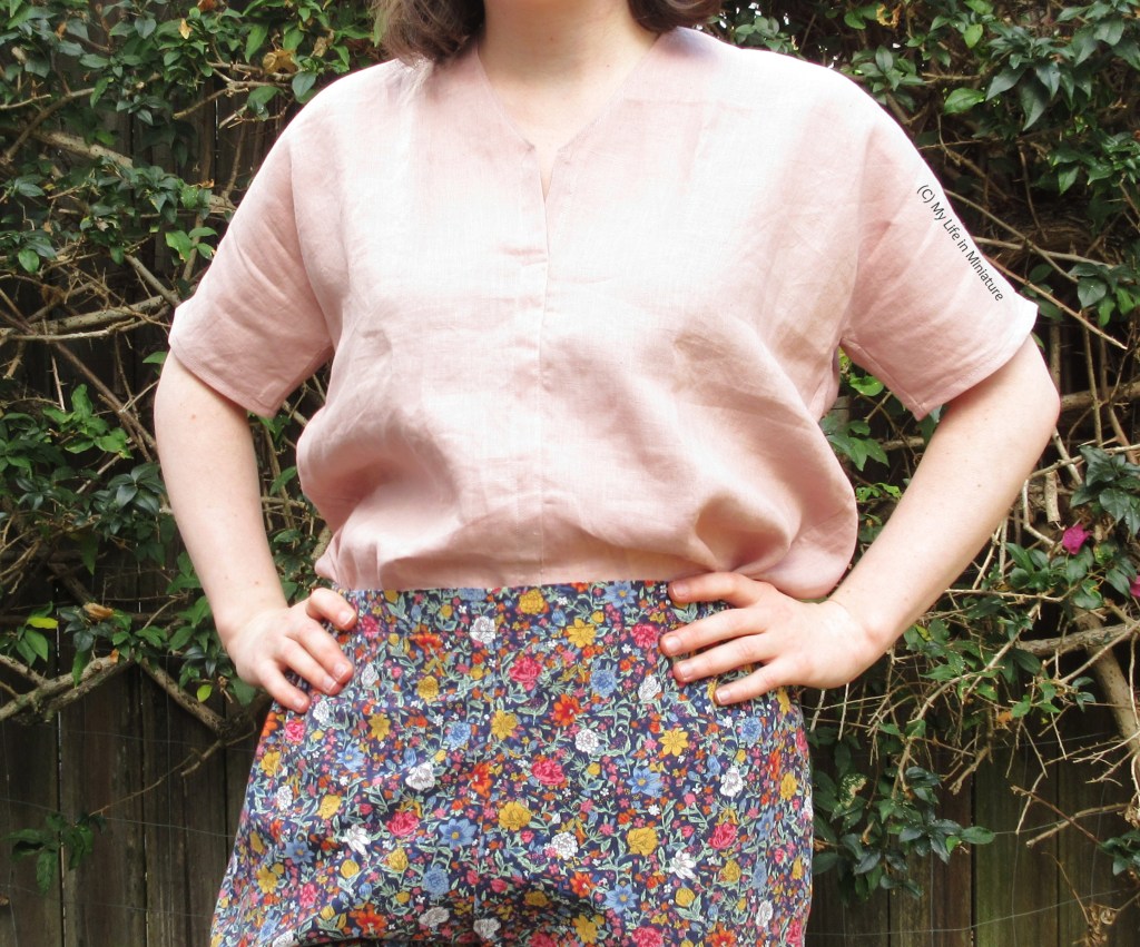 The author stands against a hedge, photographed from shoulders to thighs. She wears the same pale pink top and floral pants combination. The shirt is tucked into the pants, and her hands are on her hips. 