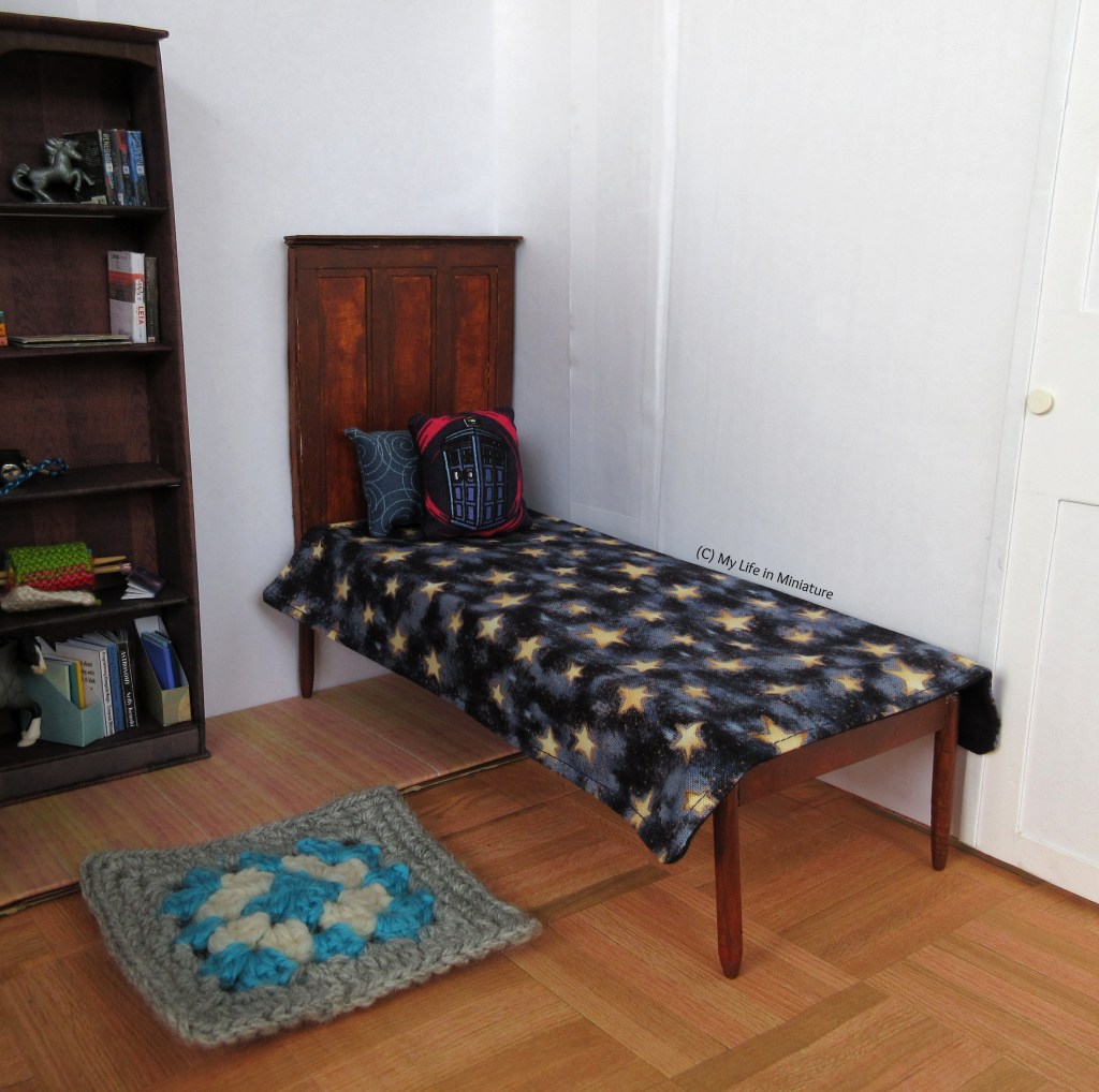 The interior of Sarah's bedroom, which has white walls and wooden floors. The bed is dark wood, with a panelled headboard. The new bedspread is navy with yellow stars on it, and the two cushions resting against the headboard are also navy. There is a blue, white, and grey rug on the floor, and Sarah's bookshelf can be seen to the left of the image. 