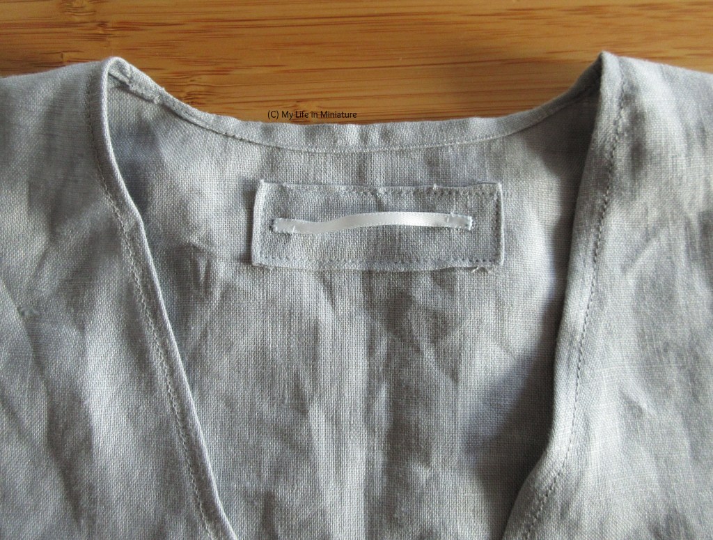 Close-up of the pale blue shirt, laid flat on a wood surface. Close-up shows the back neck area, where there is a rectangle of linen with a small piece of pale blue ribbon sewn to it.