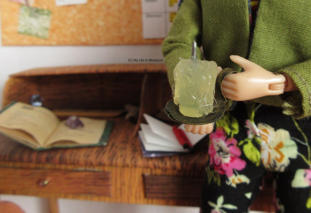 Close-up of Sarah's hands as she stands near her desk in her room. She is holding an unlit candle on a small black metal plate with a mug-like handle. Her desk, blurred in the background, is covered in open books, pencils, and crystals. 