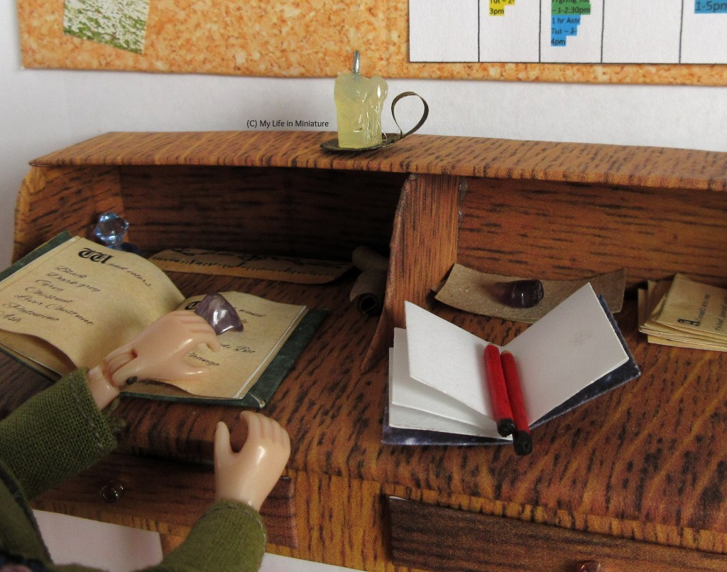 Close-up of Sarah's desk, where a candle in its holder is on the shelf above, unlit. Sarah's hands can be seen resting on the desk's surface and on an open book. Also on the desk are papers weighed down eith crystals, and an open notebook with pencils in.