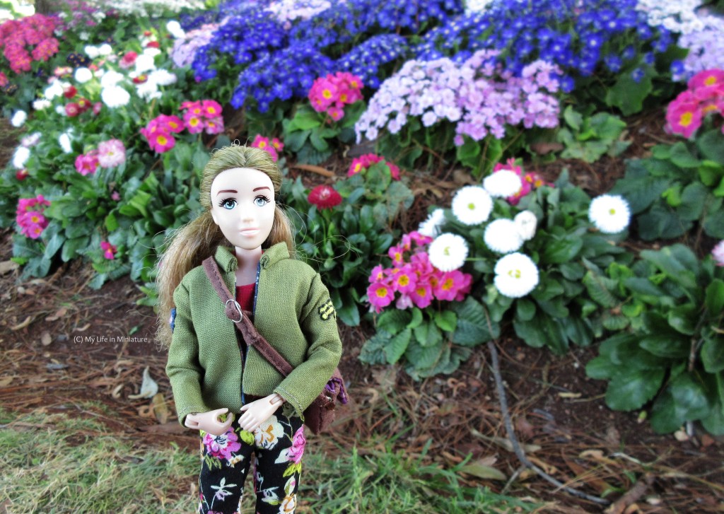 Sarah stands in front of a bed of blue, purple, pink, and white flowers. She smiles at the camera, wearing her khaki jacket with black floral pants. Her brown stachel is slung over her shoulder. 