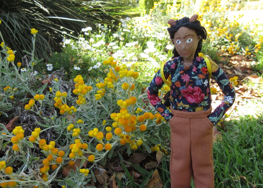 Petra stands on grass under a tree, walking towards the camera beside a garden bed. She has her hands in her pant pockets as she looks at the small yellow flowers in the bed. Her yellow backpack's straps are visible on her shoulders. 