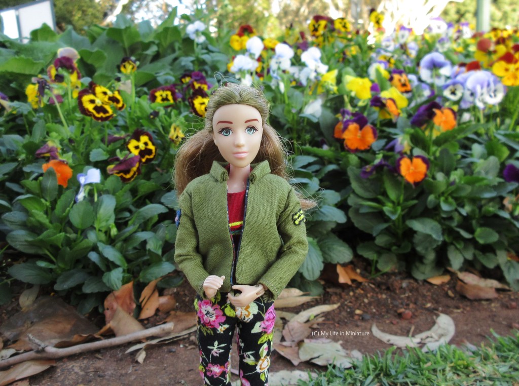Sarah stands in the shade, in front of a garden bed filled with pansies in various colours and patterns. She smiles at the camera, wearing her khaki jacket and black floral pants. 