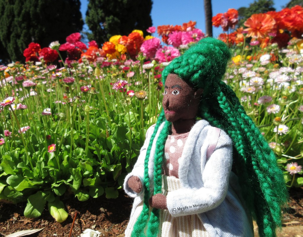 Hazel stands in front of a garden bed, filled with tall flowers in shades of pink and yellow. She wears her grey cardigan with a cream striped skirt, and is looking to the left of the shot. Some of her green braids have been pulled forward voer her shoulder. 