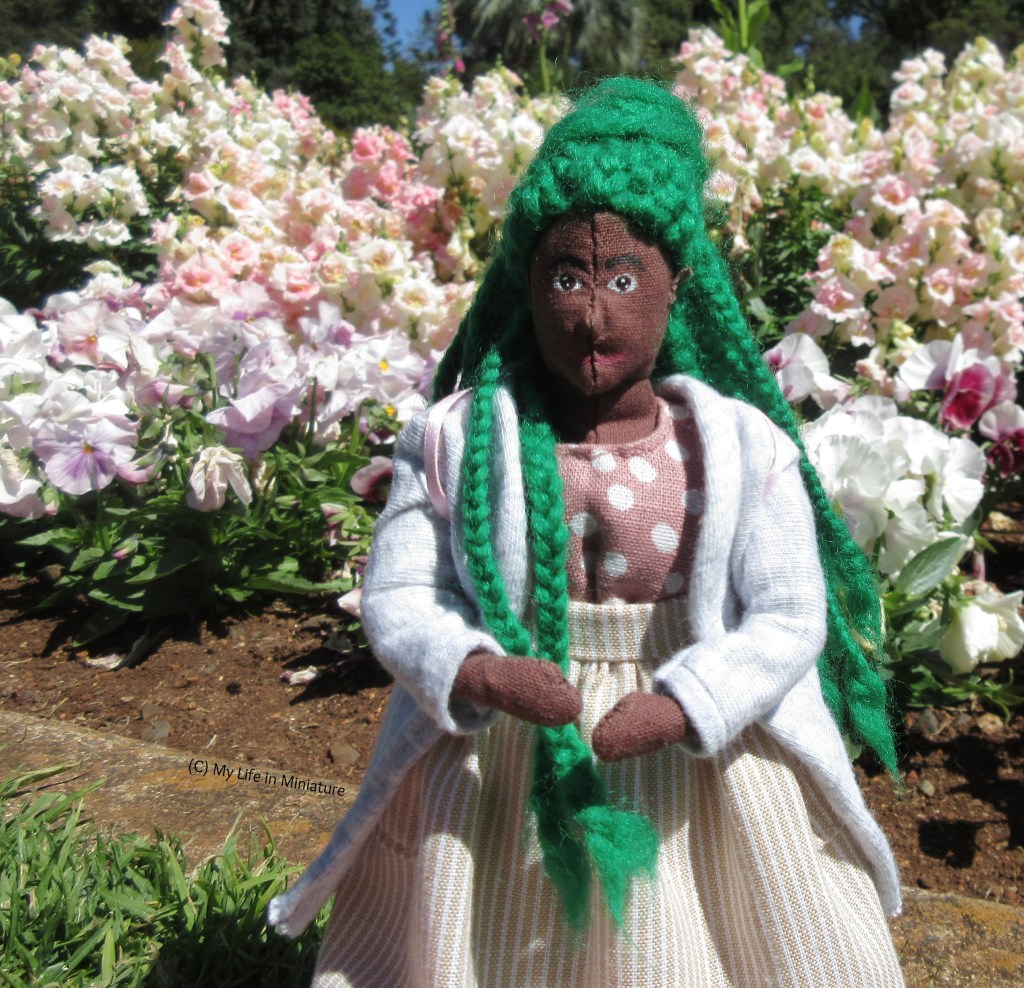 Hazel smiles at the camera, standing in front of a bed of white, pale purple, and pale pink flowers. She wears her grey cardigan over her cream-striped skirt and pink polka-dot dress. Two of her green braids are pulled over one shoulder. 