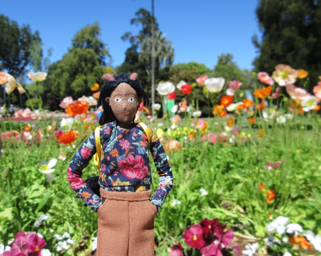 Petra stands in front of a sunny garden bed, smiling at the camera with hands in her pockets. The bed is filled with tall poppies in shades of red, orange, pink, and white. 