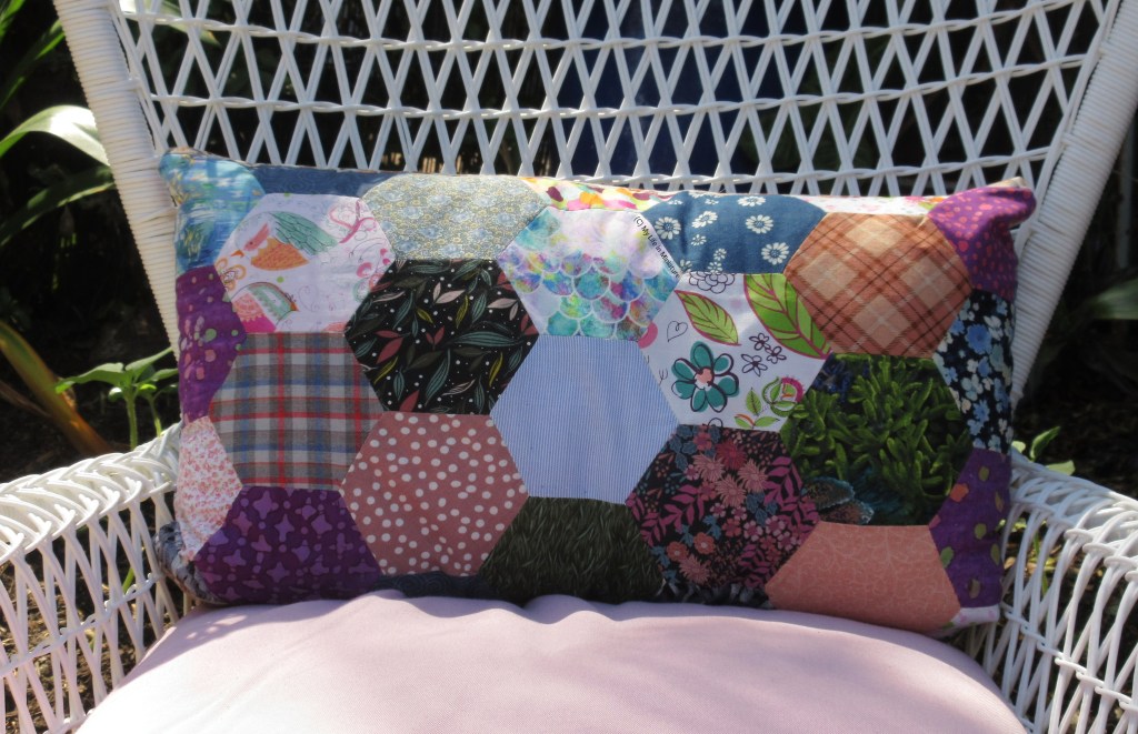 A rectangular cushion is resting against the back of a white outdoor chair, in the shade of a tree. The cushion cover is a patchwork of many different fabrics in all different colours; florals, stripes, checks, abstract designs. 