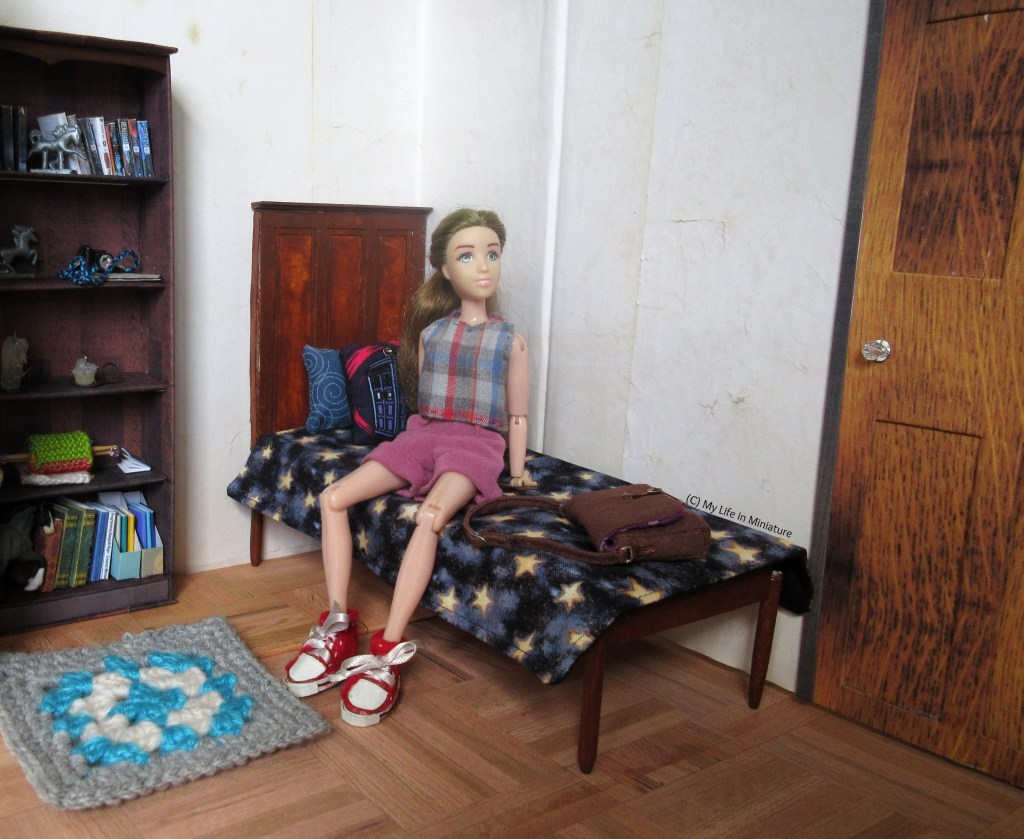 Sarah sits on her bed in her bedroom, looking around. She wears pink corduroy shorts with a blue checked shirt and red shoes. The walls are the same tea-dyed cream as before. To the right of the bed is her bedroom door, which is now a darker wood with a white bead as a doorknob.