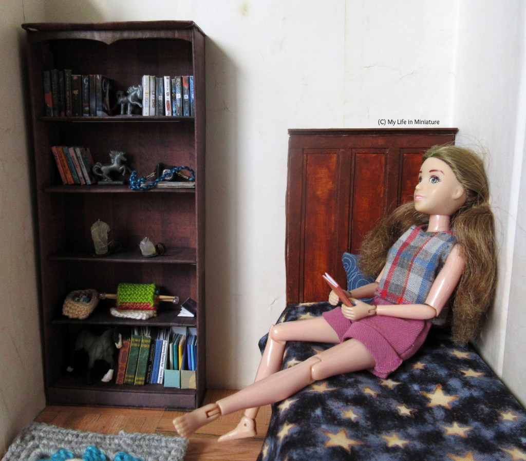 Sarah sits on her bed, leaning against the wall with her legs tucked up beneath her. She has her shoes off, and a book in her lap. Her hair is out of its ponytail, loose behind her back. 