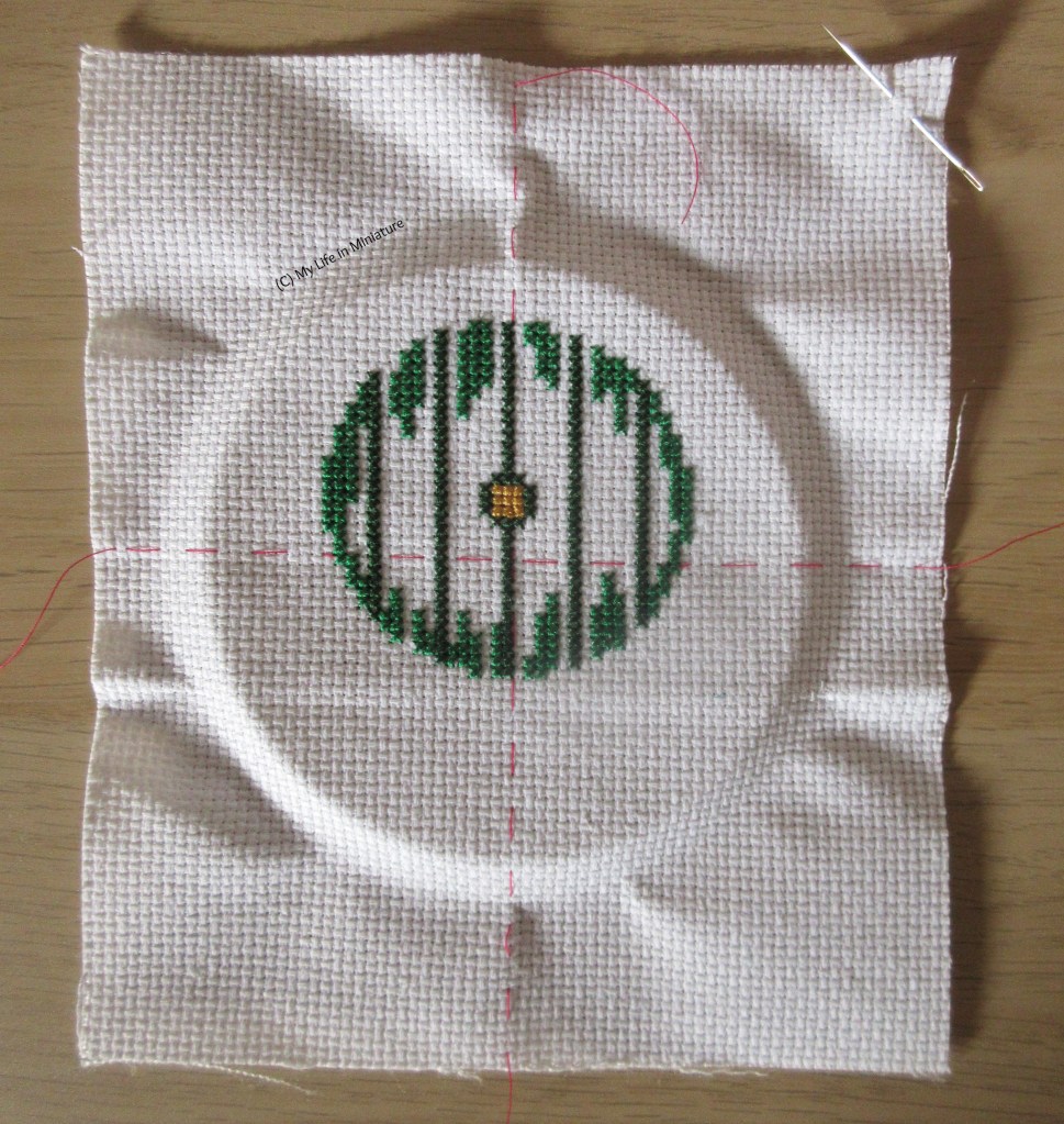A piece of white Aida cloth is flat on a wood surface. It has a needle pushed through the top right-hand corner, and some cross stitching in the middle. The stitching is in various shades of green, and is in a rough circle. 