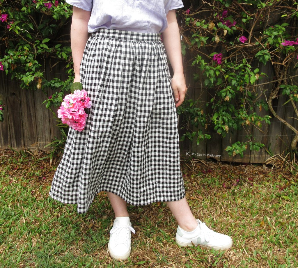 The author stands against the bougainvillea bush, wearing the gingham skirt. The skirt is blowing against her legs, and she has one foot pointing to the right, as if she's about to walk that direction. Her hands hang at her sides, and she's holding a bouquet of fake pink flowers in one hand.