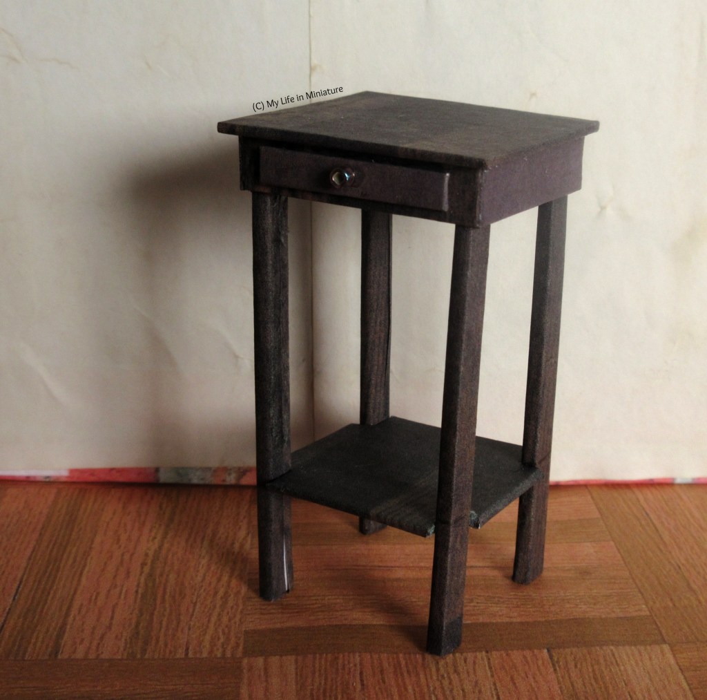 Close-up of the bedside table. It's dark wood, roughly square, with a drawer underneath the top surface and an additional shelf near the bottom of the four square legs. It's photographed on its own against a cream wall, on wood floors. 