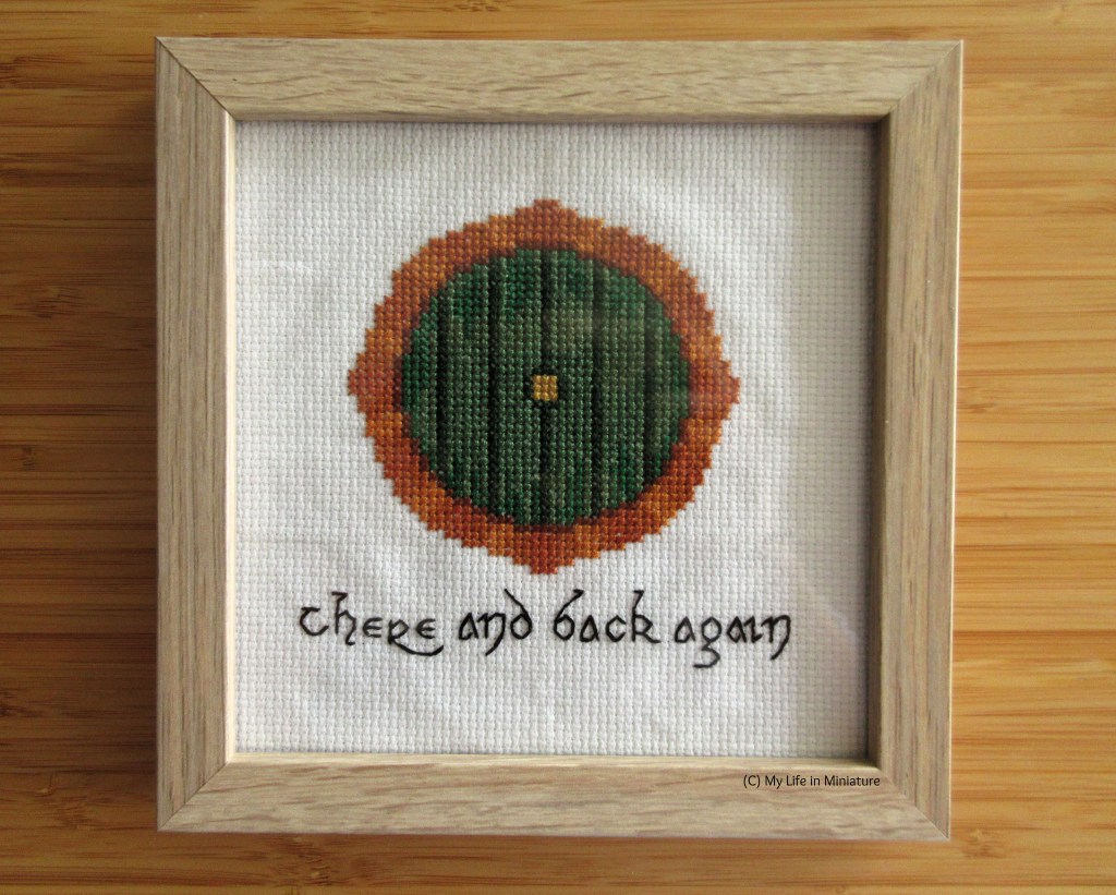 A framed cross-stitching sits flat on a wood surface. It is of a Hobbit-hole door, green and circular with bricks around it. The black text beneath the door says 'there and back again', in an Elvish-esque font. The frame is square and wooden, very simple in style. 