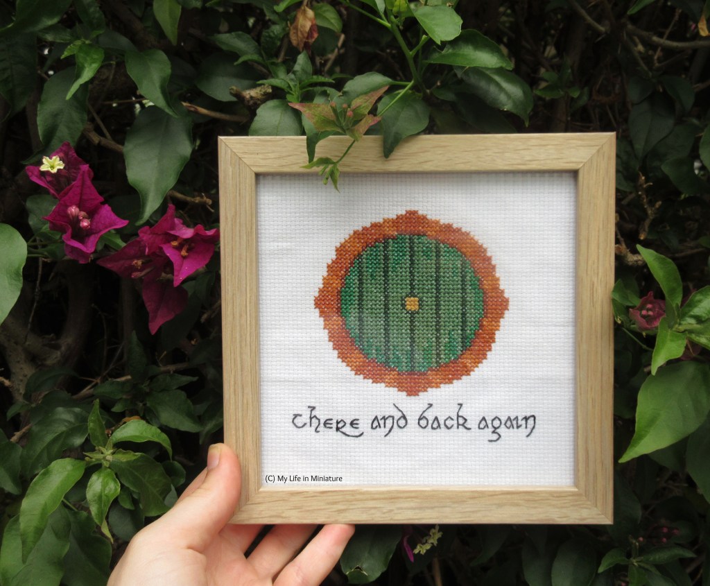 The author's hand holds the framed Hobbit-hole stitching up to a hedge, where some leaves curl over the frame and pink flowers are visible beside the stitching. 