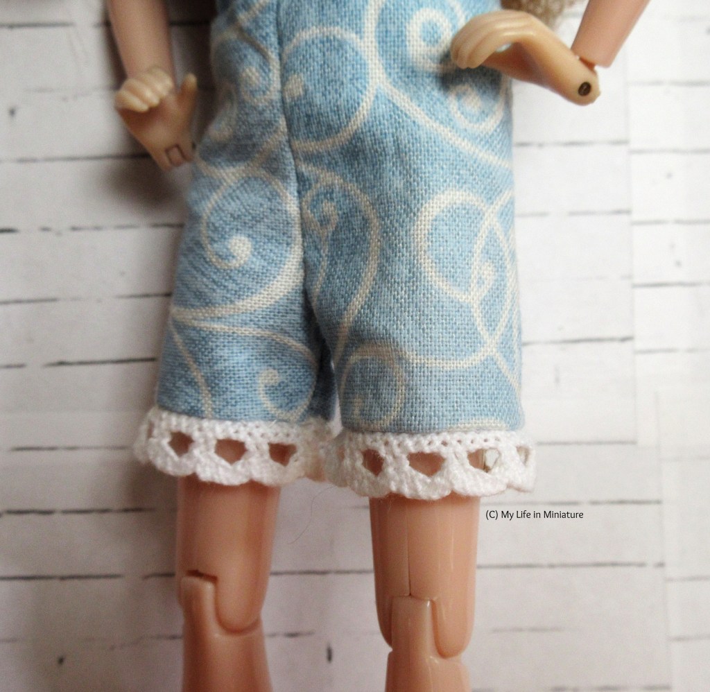 Close-up of the white lace hem of Sarah's playsuit. The lace falls slightly above her knees, where before it was covering her knees. 