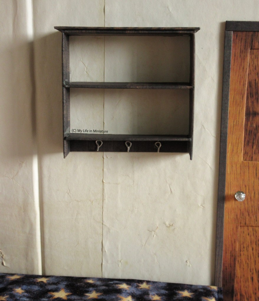 A brown wood wall-mounted shelving unit is on the cream wall above Sarah's bed, and to the left of her bedroom door. It has two shelves, and there are three silver hooks underneath the bottom shelf.