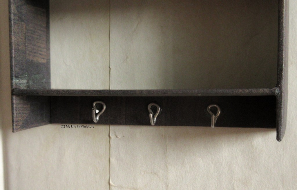 Close-up of the hooks under the lower shelf. They're silver, made of wire bent into shape. They are mounted onto a wooden board between the bottom of the sides and the bottom shelf, that sits flush against the wall.