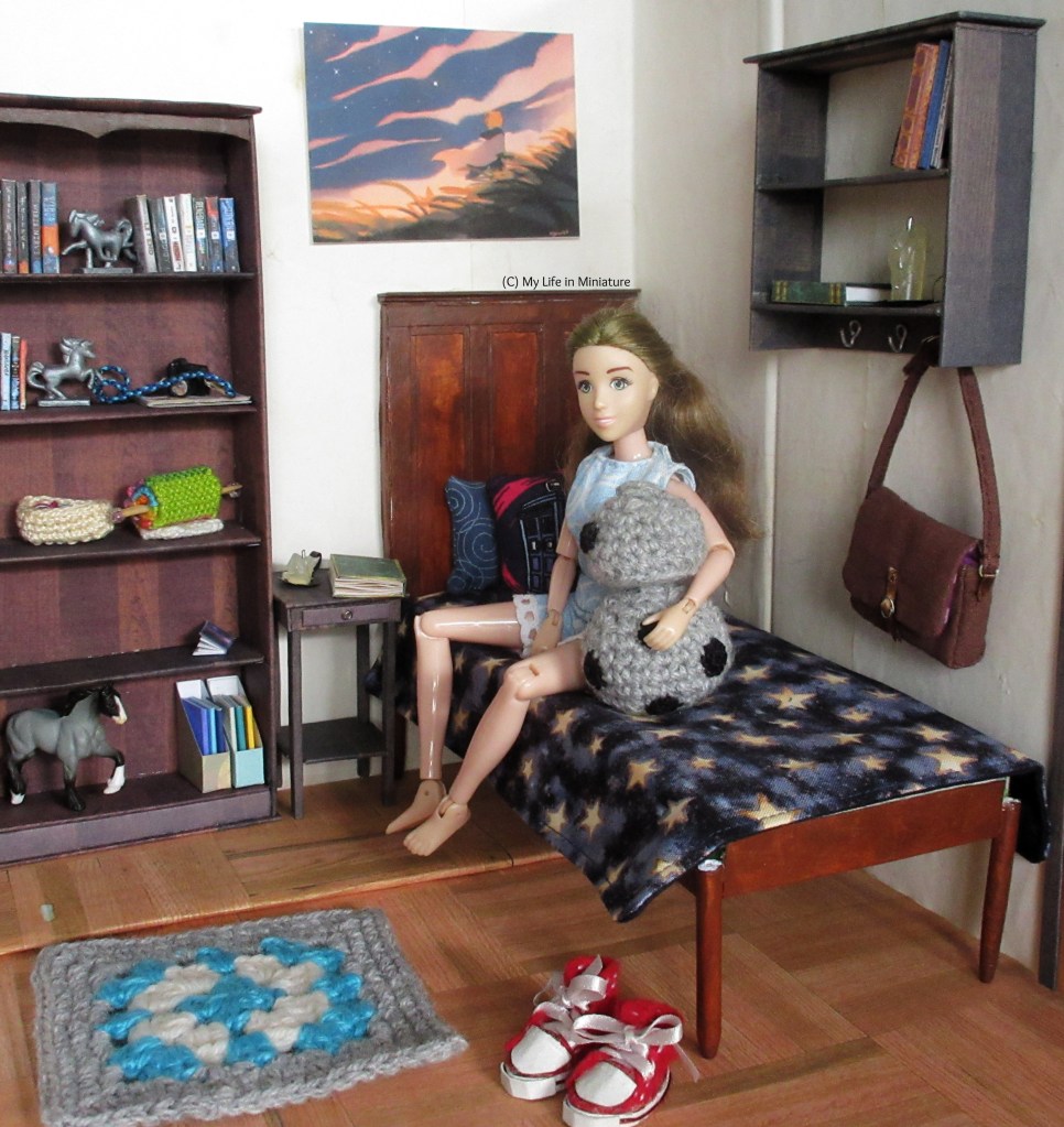 Sarah sits on her bed beside Chip the android, with one arm around them. The textbook is on the bedside table, with a candle. The rest of the room is lightly cluttered with books, candles, statues, a knitting project, and cameras. Sarah's red shoes are on the floor near the base of the bed. 