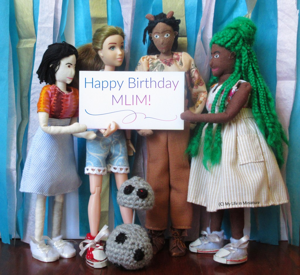 Tiffany, Sarah, Petra, and Hazel stand against a backdrop of streamers, with Chip the android at their feet. Chip's head is elevated, looking directly at the camera. The four girls hold a sign between them that says 'Happy Birthday MLIM!'. They're all looking at the sign and/or smiling at each other.