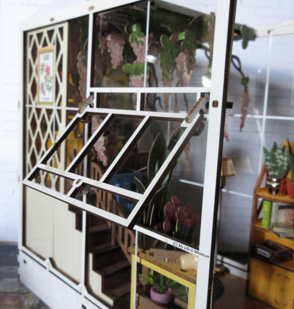 Close-up of the side of the exterior of the book nook. Behind one of the small tables near the entrance is a section of the greenhouse that angles outward; an openable window. This window is shown from the outside.